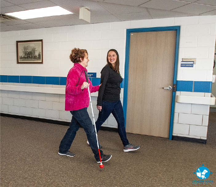 A person who is blind, using a white cane, receiving human guide services in a hallway. Photo provided by the Leader Dogs for the Blind. 