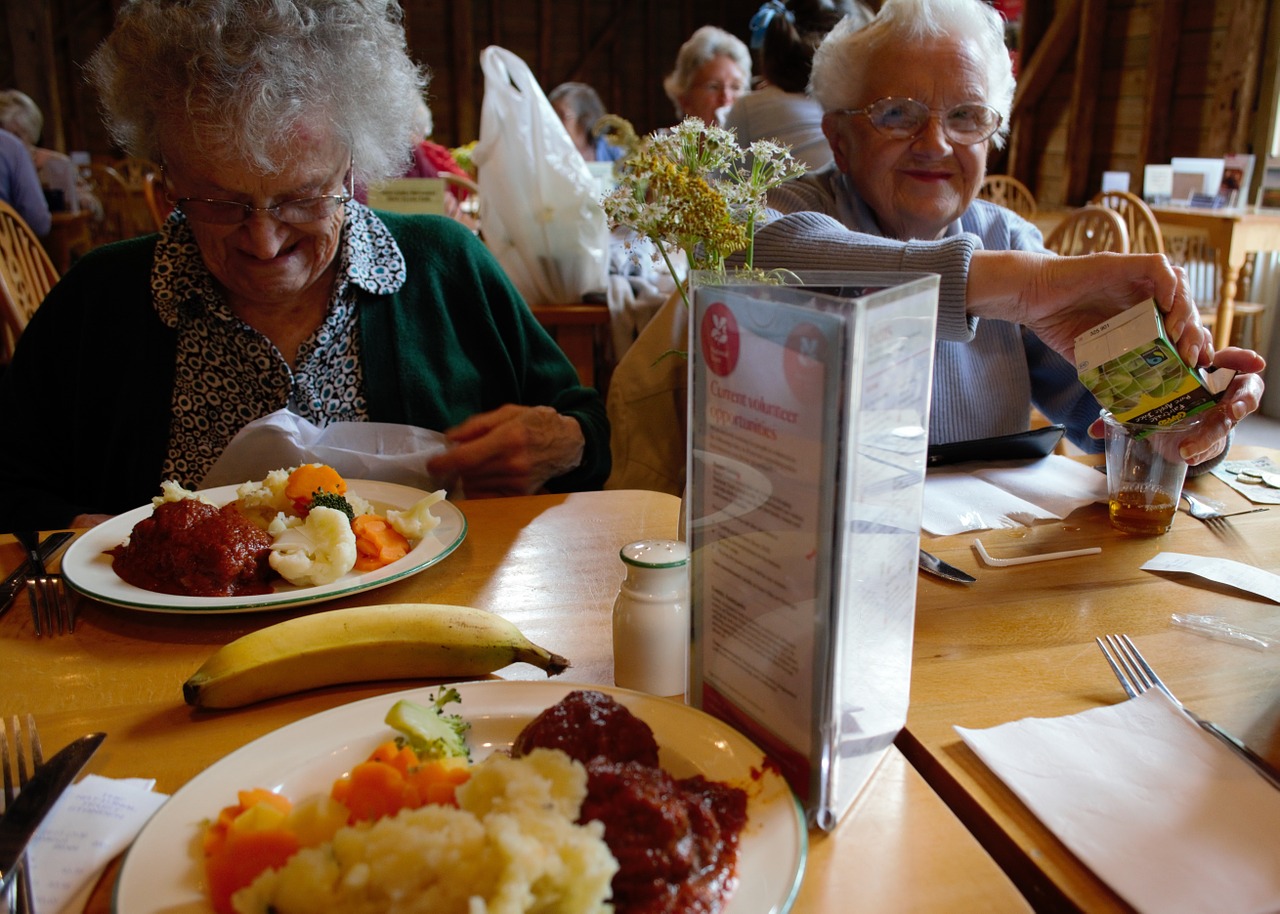 two older white women with grey and white hair sitting next to each other at restaurant eating dinner smiling